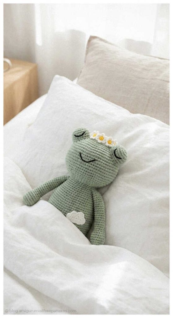 Soft green amigurumi frog doll lying on white bedding with small daisy detail, free crochet frog pattern idea