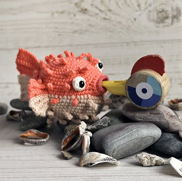 Cute amigurumi puffer fish crochet toy on beach stones with seashells, free crochet fish pattern