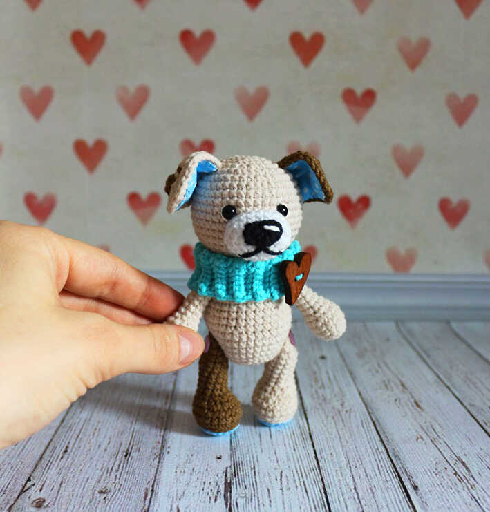Amigurumi puppy dog with blue scarf and heart detail held by hand on wooden floor, free crochet dog pattern