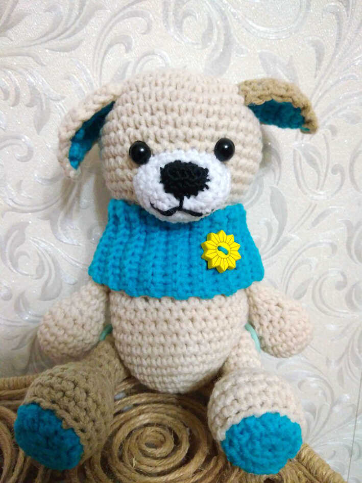 Amigurumi puppy dog wearing a blue sweater with flower detail sitting on rope surface, free crochet dog pattern