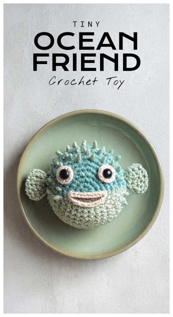 Tiny blue amigurumi puffer fish crochet toy sitting on a plate, adorable handmade sea creature plush
