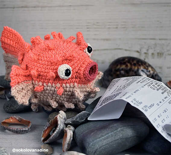 Cute amigurumi puffer fish crochet toy sitting on beach stones with seashells, free crochet fish pattern