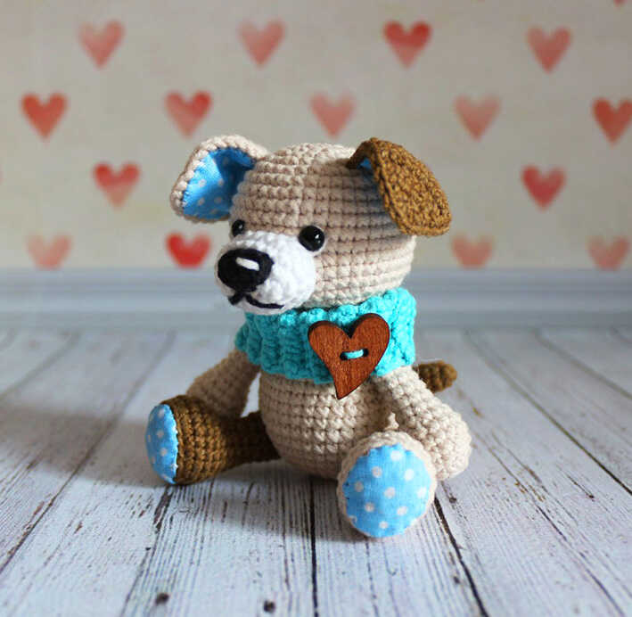 Cute amigurumi puppy dog with blue scarf and heart detail sitting on wooden floor, free crochet dog pattern