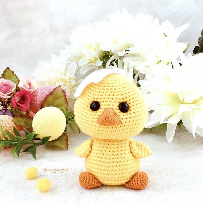Cute yellow amigurumi chick with orange beak and feet sitting in front of flowers, free crochet chick pattern