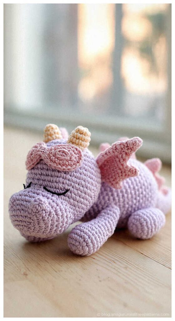 Sleeping pink amigurumi dragon plush resting on a wooden table, cute crochet dragon toy idea