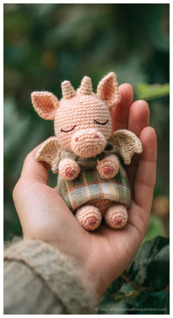 Sleeping green amigurumi dragon sitting by a window wearing a small scarf, cute crochet dragon plush toy idea