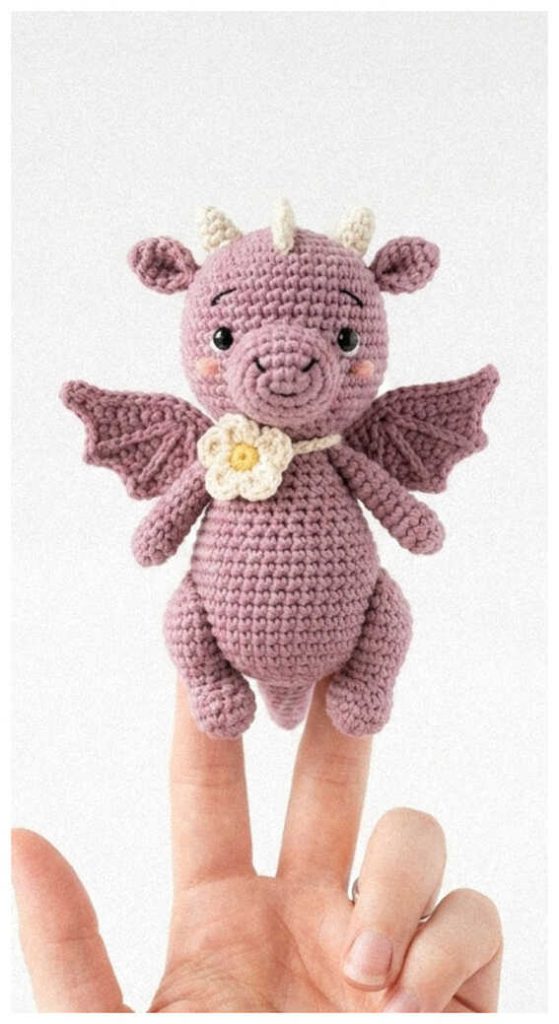 Small pink amigurumi dragon with wings and a tiny flower standing on fingers, cute crochet dragon plush toy