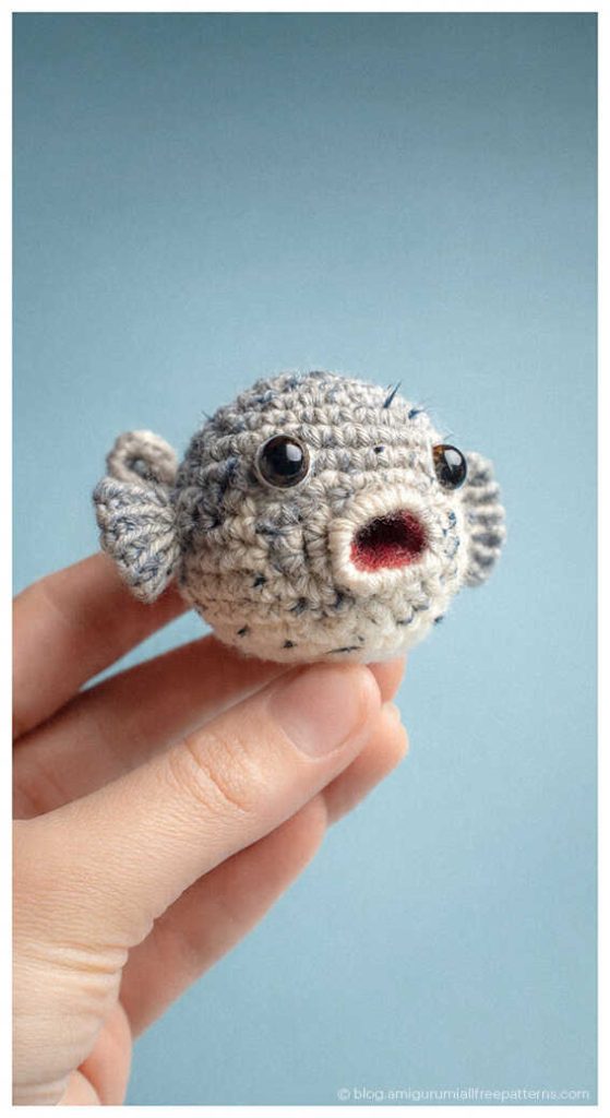 Small gray amigurumi puffer fish crochet toy held between fingers, cute handmade ocean plush idea